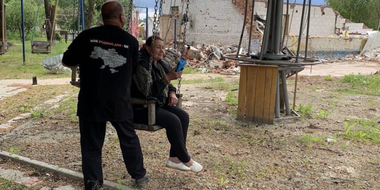 Nataliia Pasiuha, holding the Chuyka drone detector, sits on a swing in the frontline village of Spirne. Behind her lie the ruins of the café that served as a distribution point for Vorskla